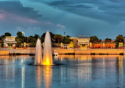 Waterfront scene featuring a lit up double fountain in the foreground, with a building labeled "Cantina" and a bridge in the background under a partly cloudy sky.