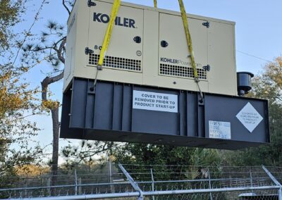 Crane hoisting a Kohler generator over a fenced area, with trees and blue sky in the background.