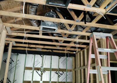 Construction site with exposed wooden framing and ventilation ducts on the ceiling. A ladder stands against a wall with partially installed insulation and drywall.