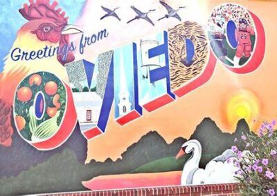 Colorful mural with "Greetings from Oviedo" in large letters, decorated with images of a rooster, oranges, sunset, a swan, and flying birds.