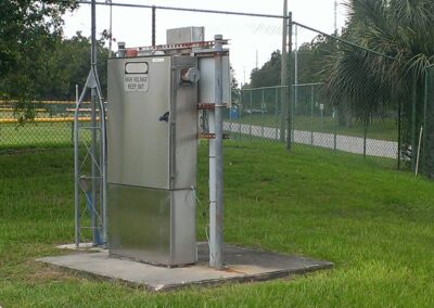 Metal electrical box labeled "High Voltage Keep Out," situated on a concrete slab near a grassy area with a chain-link fence and palm tree in the background.