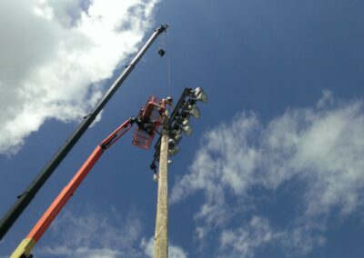 A crane lifts a worker in a red basket to repair or inspect a tall light pole under a clear blue sky with clouds.