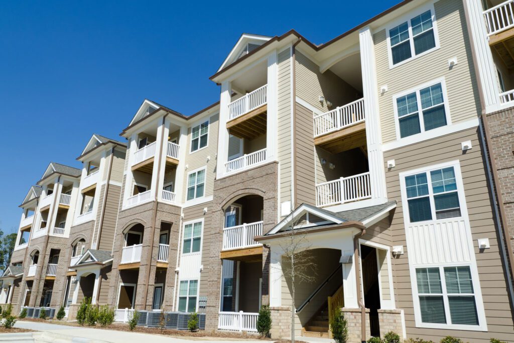 Three-story apartment building with balconies and beige siding, set against a clear blue sky.