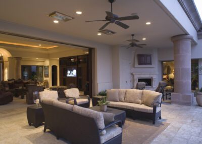 Spacious covered outdoor patio with wicker furniture set, ceiling fans, and potted plants. The area opens to an indoor living room with plush seating and a fireplace.