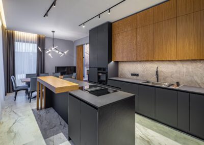 Modern kitchen with black cabinets, wood accents, and marble floors. Contains a central island, built-in oven, cooktop, and geometric chandelier. Dining table and chairs are visible in the background.