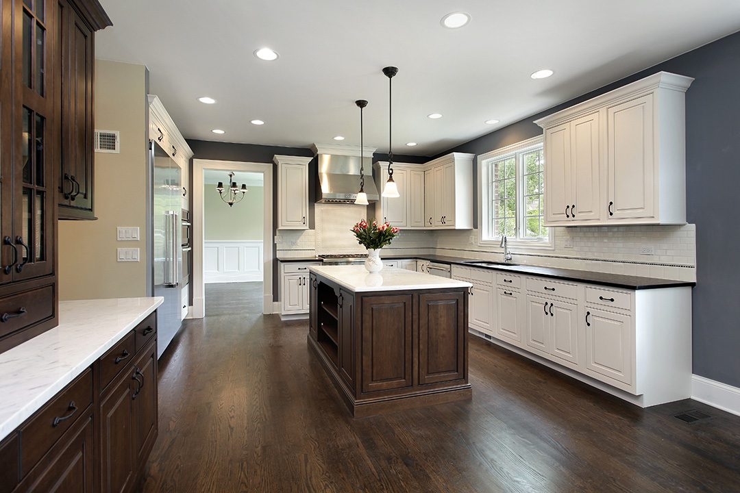 Spacious kitchen with white cabinetry, dark island, pendant lights, and hardwood floor. Large window above sink, leading to adjacent dining area with visible chandelier.
